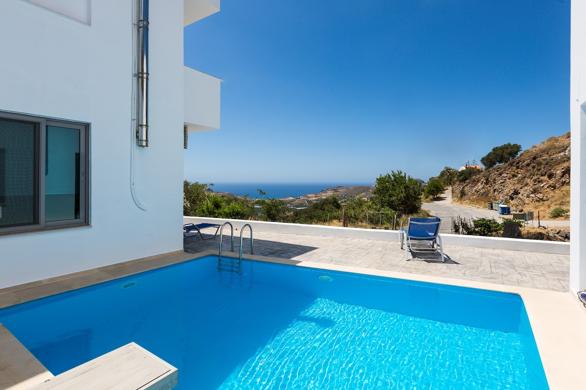 A bright swimming pool is centrally positioned with inviting blue water reflecting the clear sky. Lush greenery and a scenic landscape with distant sea views surround the pool area. A lounge chair is visible on the patio, encouraging relaxation in the sun.