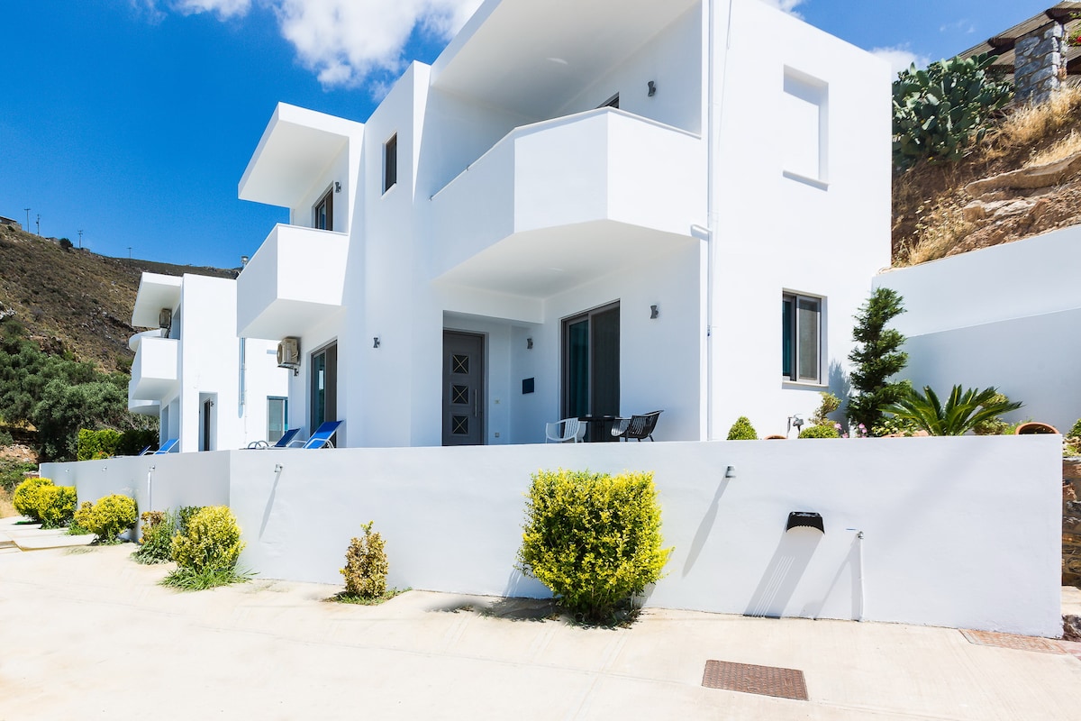A modern complex of three identical maisonettes is presented, showcasing a clean white exterior. Each maisonette features a balcony and large windows. Neat landscaping with low shrubs and a few palm trees can be seen, along with a clear blue sky above.