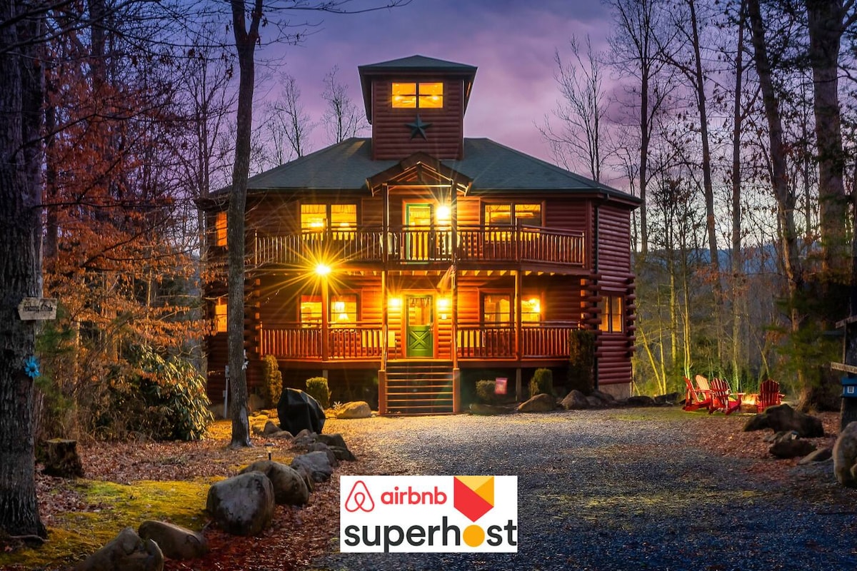 A spacious log cabin is surrounded by trees during twilight. The structure features multiple levels with large balconies, warm lights illuminating the exterior. A gravel drive leads to the entrance, lined with natural stone and outdoor seating. Tranquil surroundings enhance the cabin's secluded charm.