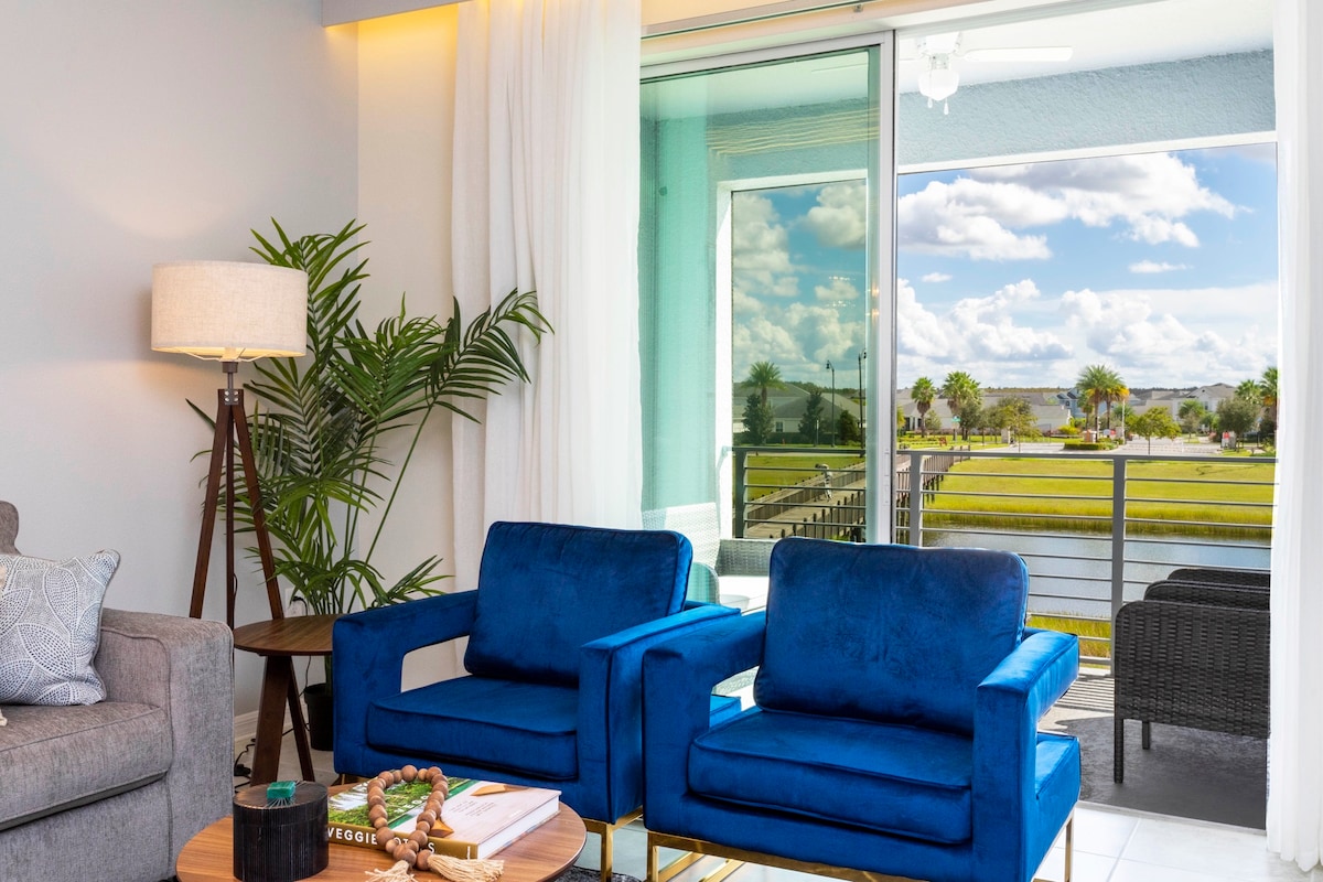 A bright living area features two blue armchairs, a wooden coffee table adorned with decor, and a plush gray sofa. Floor-to-ceiling windows offer a view of the outdoors, with palm trees visible outside, allowing natural light to fill the space.