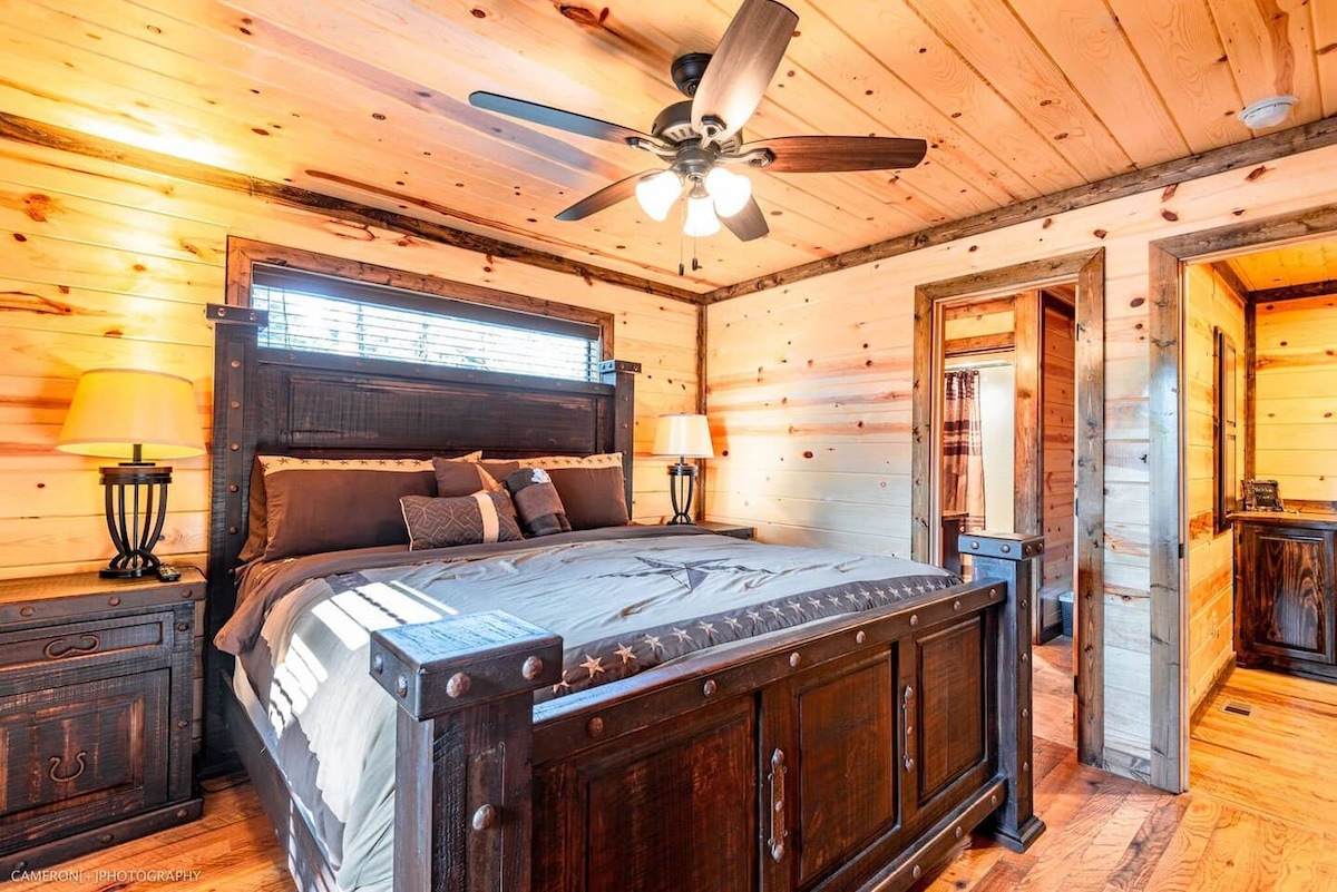 Spacious main-level bedroom featuring a luxurious king bed, ensuite bathroom, rustic wood furnishings, and ceiling fan-perfect for restful nights and ultimate comfort during your cabin getaway.