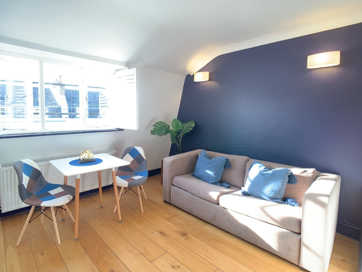 Natural light floods a cozy living area, where a comfortable sofa is paired with a small dining table for two. A simple plant adds a touch of greenery against a stylish dark blue wall, and warm wooden flooring enhances the inviting ambiance.
