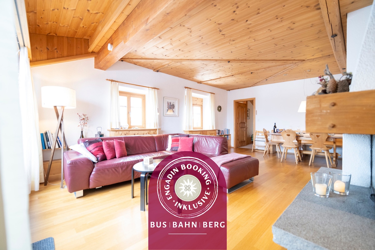 A spacious living area features wooden beams overhead and large windows allowing natural light to fill the space. A leather sofa is arranged with accent pillows, while a wooden dining table is visible in the background, accompanied by several chairs.