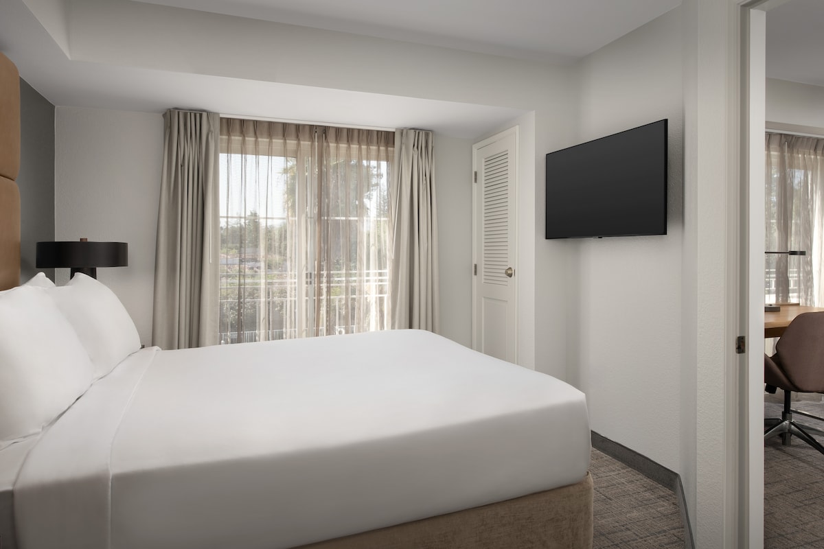 An air-conditioned room features a spacious bed with crisp white linens. Natural light filters through sheer curtains, illuminating the modern decor. A wall-mounted TV is visible, along with a comfortable chair and desk in the corner, creating a cozy and functional atmosphere.