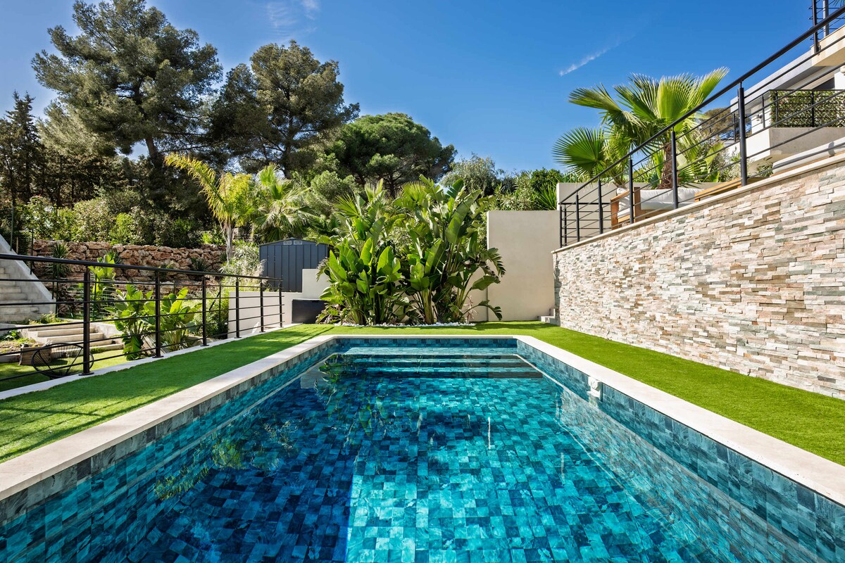 The image showcases a private swimming pool with vibrant blue tiles, reflecting sunlight. Lush greenery surrounds the pool area, including palm trees and various plants. A terraced wall with a natural stone finish provides a stylish backdrop, enhancing the outdoor space.