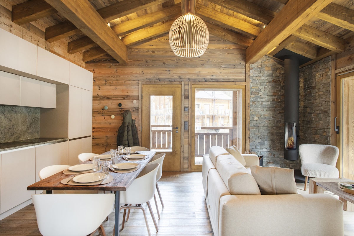 A welcoming dining area is presented with a wooden table set for six. The modern kitchen with sleek cabinetry is adjacent. Large glass doors lead to a balcony, while a cozy seating arrangement features a light-colored sofa and armchairs, complemented by wooden beams and a stone fireplace.