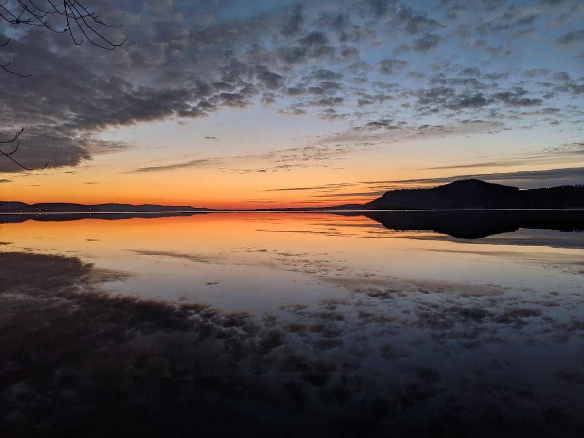 A serene lakeside view captures a vibrant sunset, with hues of orange and blue reflecting on the calm water. Clouds scatter across the sky, complementing the tranquil scene where the water mirrors the colorful sky, suggesting a peaceful natural environment.
