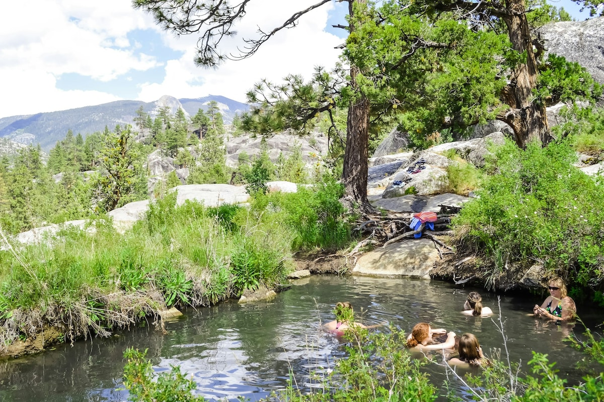 A serene natural hot spring is surrounded by lush greenery and rocky terrain. Several individuals are seen enjoying the warm water, with trees providing shade and mountains visible in the background under a partly cloudy sky.