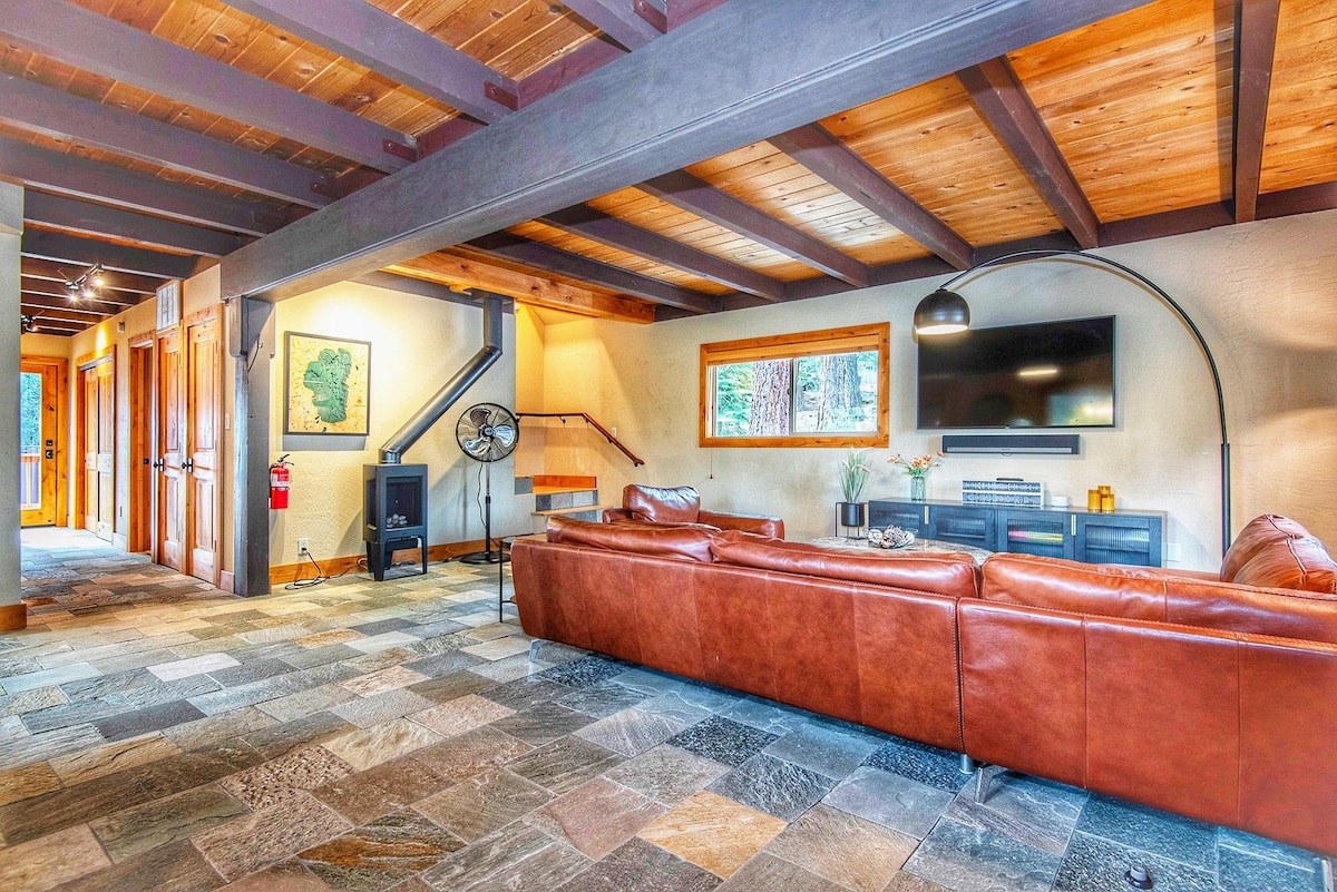 A spacious living area features a brown leather sectional sofa positioned near a wall-mounted television. Natural light is welcomed through large windows, highlighting the slate tile flooring. A staircase leads to the upper level, with additional seating and decor visible in the background.
