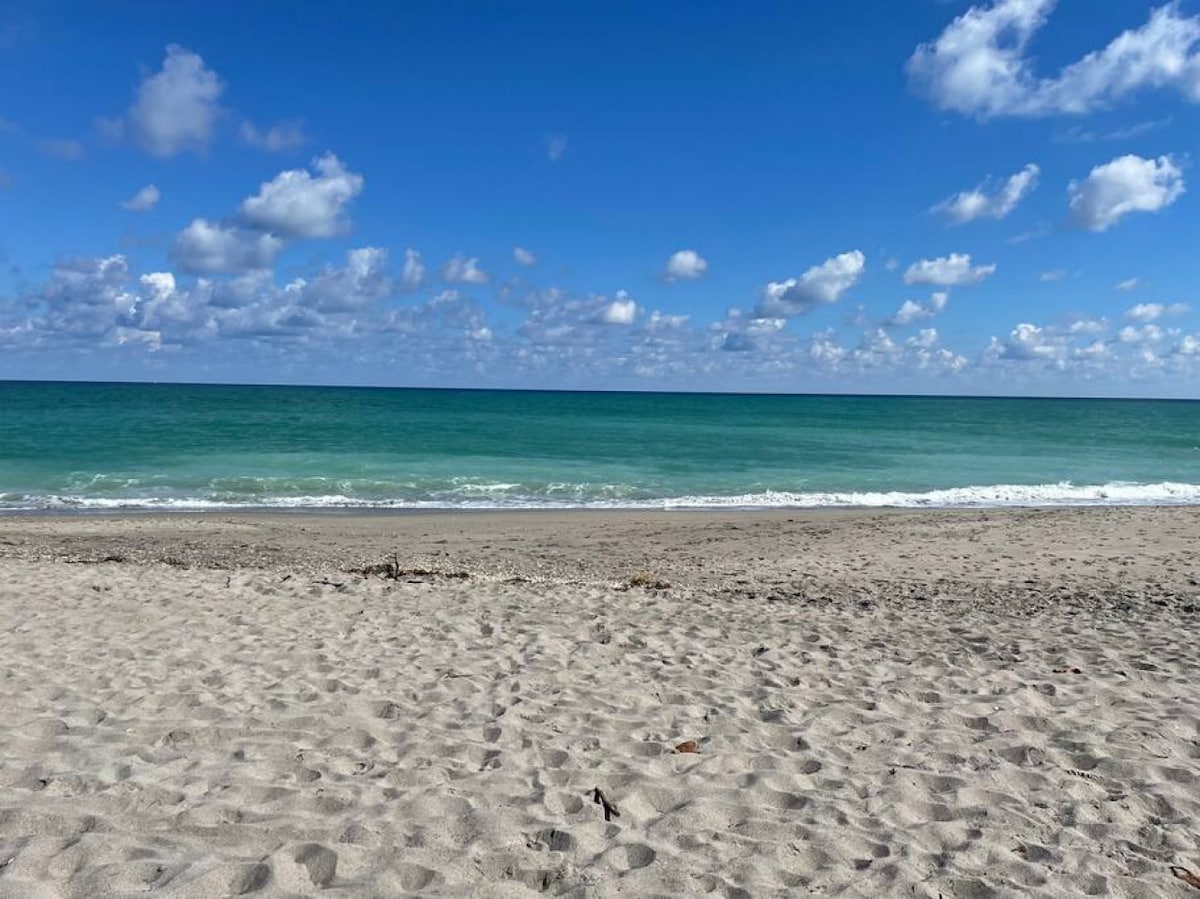 A sandy beach stretches under a bright blue sky adorned with fluffy clouds. Gentle waves roll onto the shore, creating a serene coastal view. The clear, turquoise water contrasts beautifully with the soft, light-colored sand.