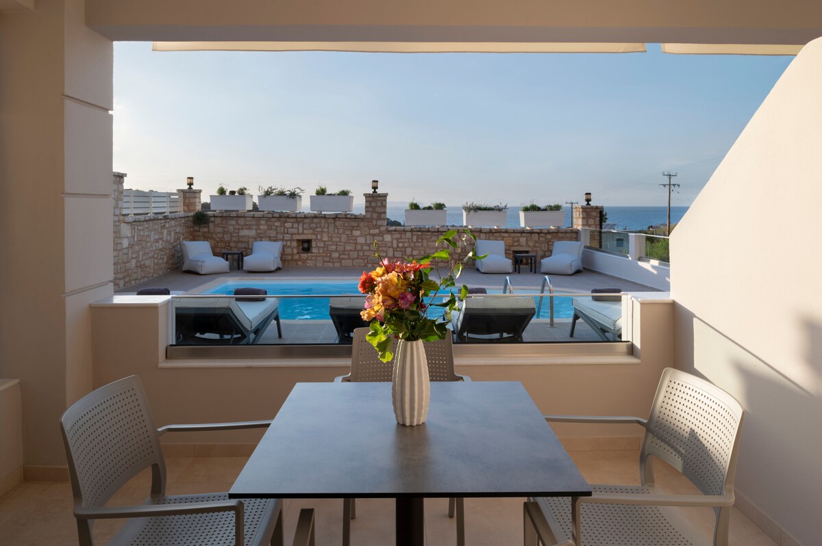 A dining area features a simple table with four chairs and a vase of fresh flowers. Beyond, a shared swimming pool is visible, surrounded by sun loungers. Clear blue skies and the distant sea create a serene backdrop.