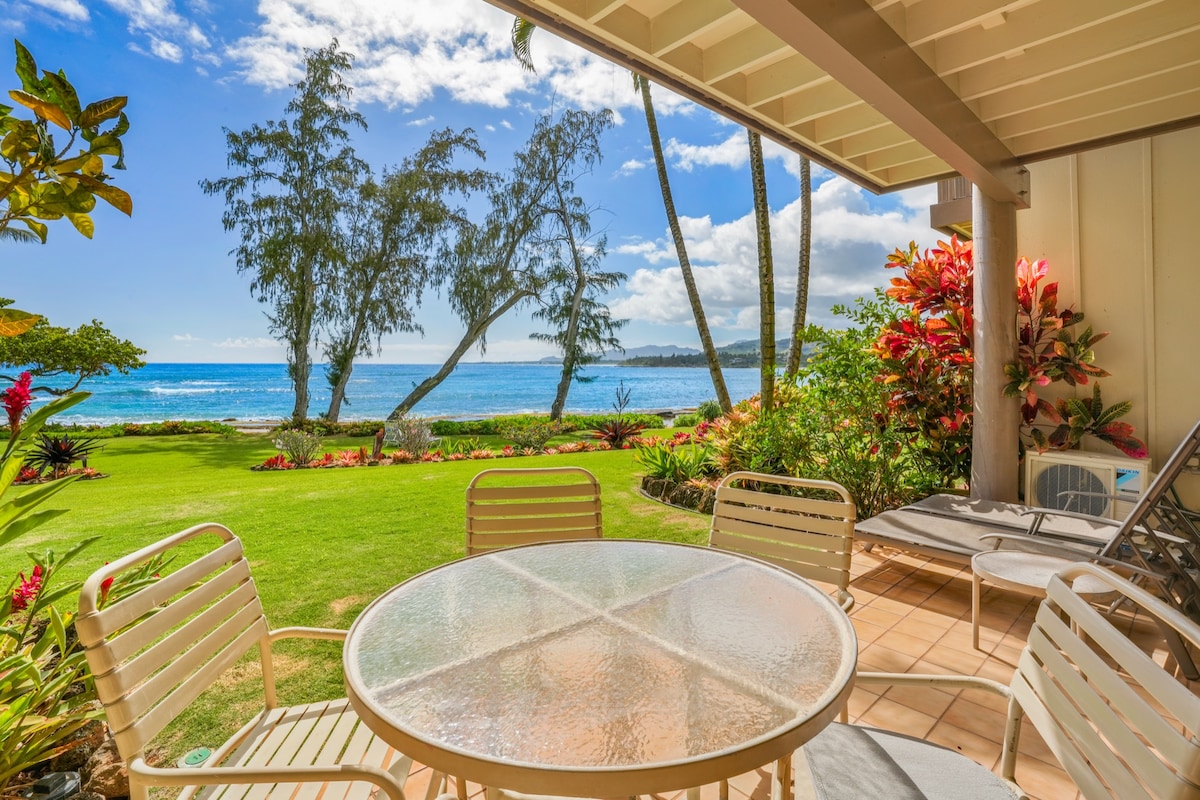 An outdoor seating area features a glass-top table surrounded by several chairs. Lush greenery and colorful tropical plants frame the view of the ocean and distant shoreline, with palm trees swaying gently under a clear blue sky.
