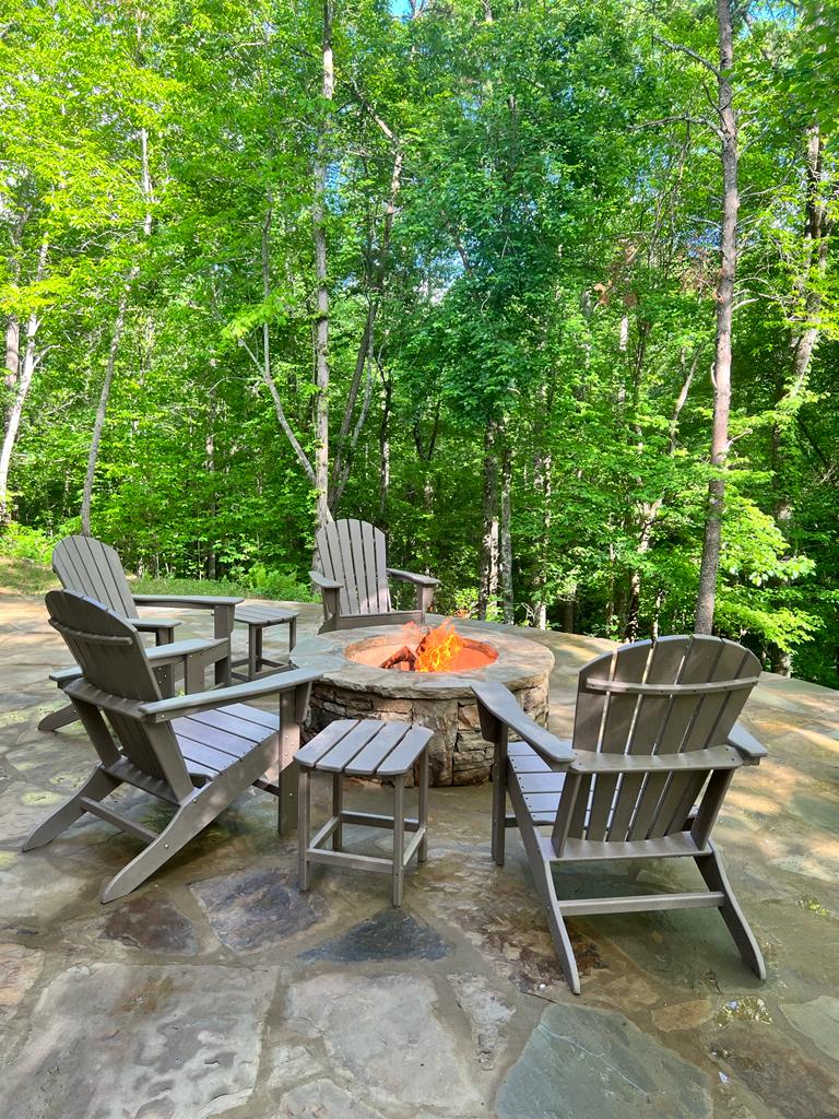 A stone fire pit is surrounded by several wooden adirondack chairs on a flagstone patio. Lush greenery from surrounding trees frames the area, creating a serene outdoor setting for gatherings. A small side table is positioned near the chairs, inviting relaxation.