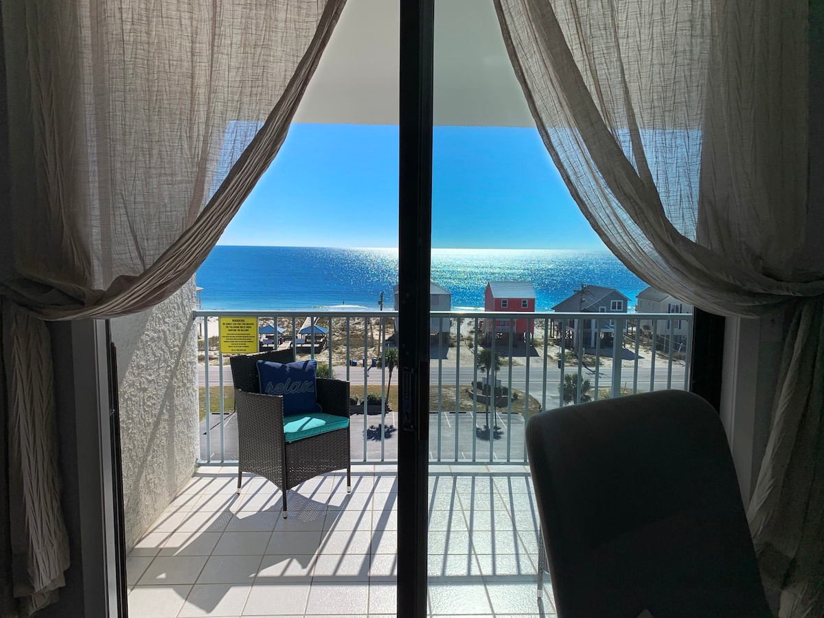 A balcony view showcases the sparkling blue ocean under clear skies. Soft curtains frame the scene, which includes a cozy chair positioned to enjoy the tranquil setting and distant beach houses. The expansive view highlights the beauty of the coastal landscape.