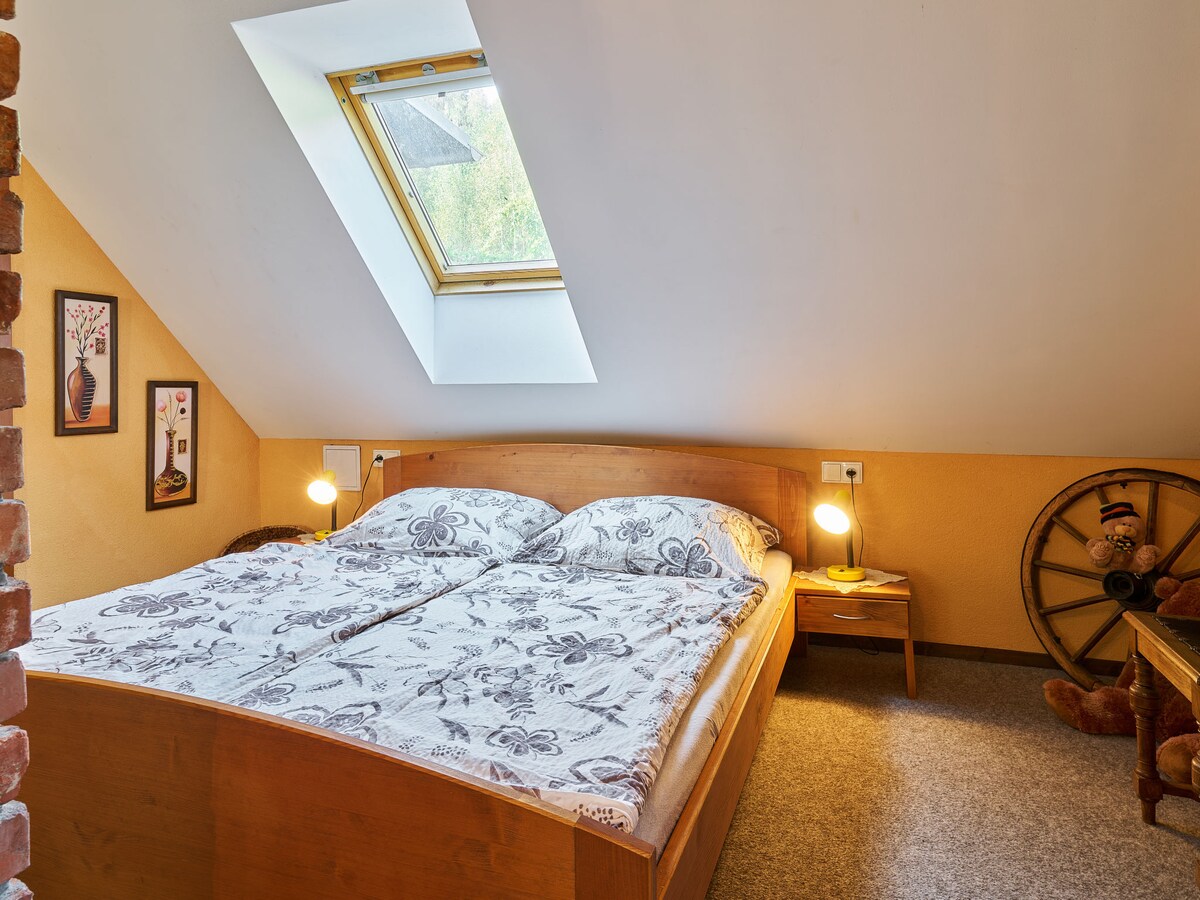 A bright sleeping area features a spacious double bed with a floral-patterned duvet. A sloped ceiling with a skylight allows natural light to enter. Nightstands with lamps are placed on each side of the bed, and decorative art pieces are displayed on the walls.
