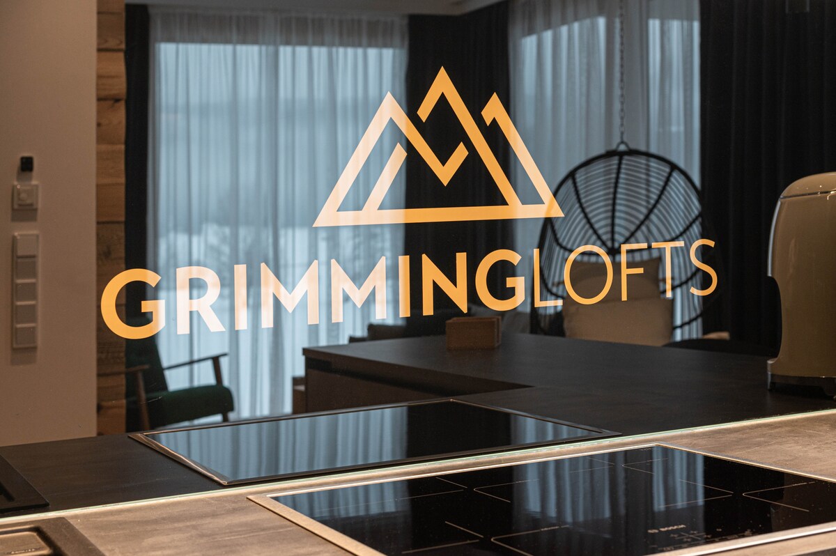 The image captures a modern kitchen space featuring a sleek countertop with an induction cooktop. A logo reading 'GRIMMINGLOFTS' is prominently displayed, while large windows are adorned with light curtains, allowing natural light to fill the area.