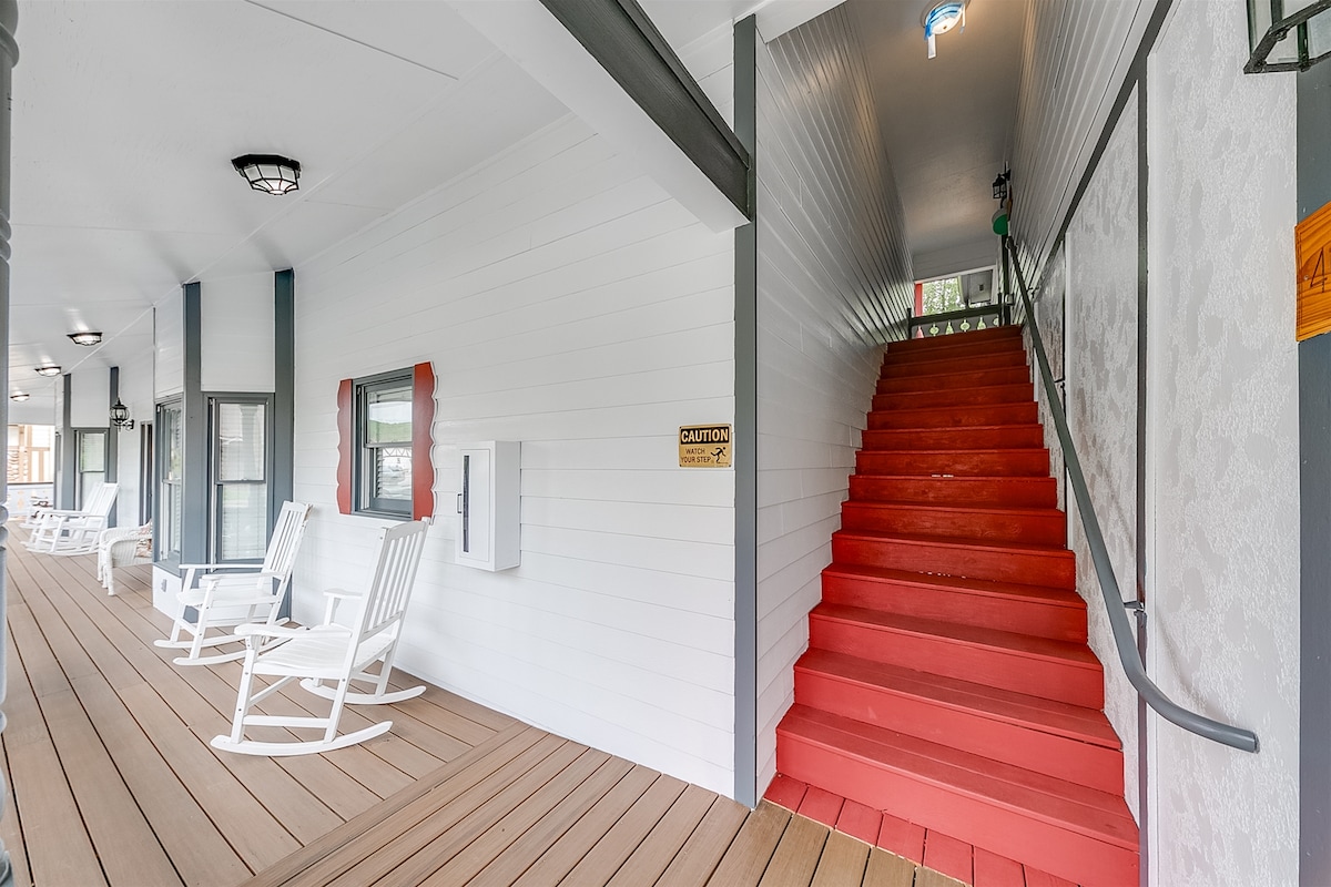 A welcoming entryway is shown with a covered porch featuring several white rocking chairs. Bright red stairs lead to the second-level unit, flanked by light-colored walls and a railing. Windows allow natural light to enhance the space.