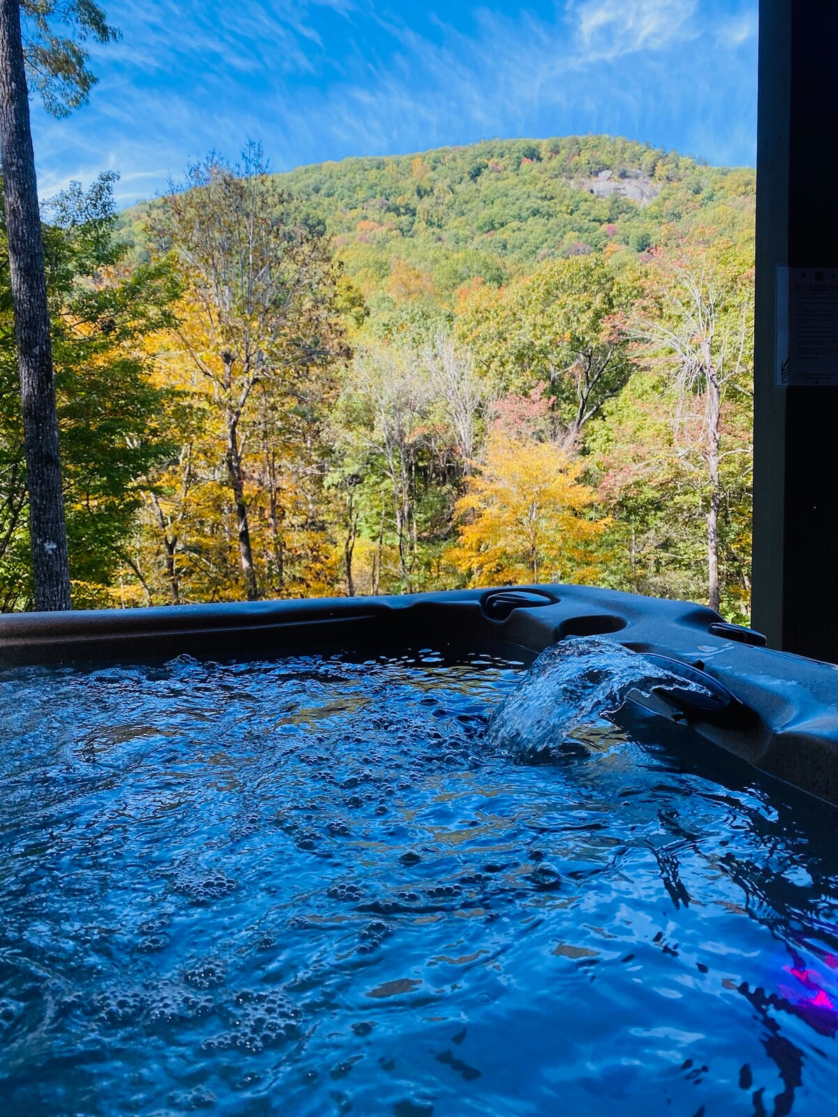 A hot tub is positioned outdoors with water gently cascading over its edge. The backdrop features lush trees and colorful autumn foliage, with distant mountains rising under a bright blue sky, creating a serene natural setting.
