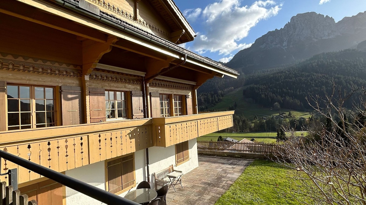 The exterior of the flat features a wooden balcony that extends along the facade, offering views of the surrounding mountains and pastures. Rugged peaks are visible in the background, complemented by expansive green fields. A stone patio area is present, along with a simple outdoor seating arrangement.