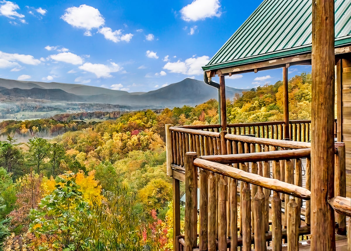 A wooden deck is outlined by rustic railings, offering panoramic views of a vibrant landscape featuring autumn foliage. Mountains rise in the distance under a clear sky, while a few clouds drift overhead, creating an expansive and serene atmosphere.