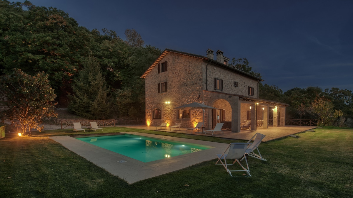 The exterior of the villa is shown at dusk, with warm lights illuminating the stone facade. A rectangular swimming pool is centered in the yard, surrounded by grass. Lounge chairs are positioned nearby, offering a comfortable space for relaxation.