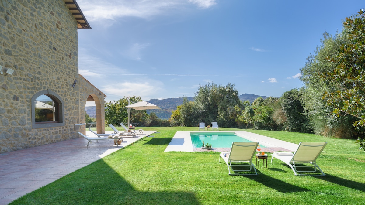 The exterior features a stone façade of the villa, complemented by landscaped green lawns surrounding a rectangular private pool. Sun loungers with umbrellas are placed beside the pool, offering a serene setting with distant hills and trees visible in the background under a clear blue sky.