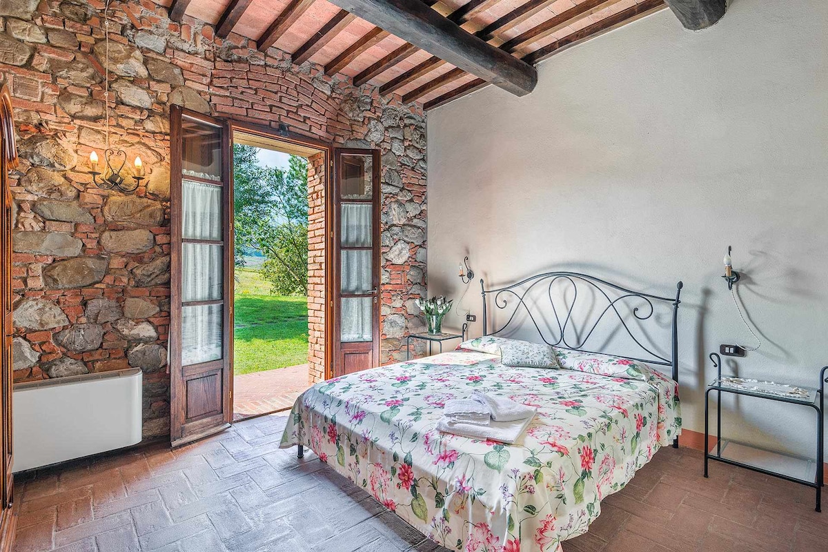 A spacious bedroom features a wrought iron bed adorned with a floral bedspread. Natural light enters through double doors that open to a scenic view. Exposed wooden beams and stone walls add rustic charm. A small metal bedside table completes the room's furnishings.