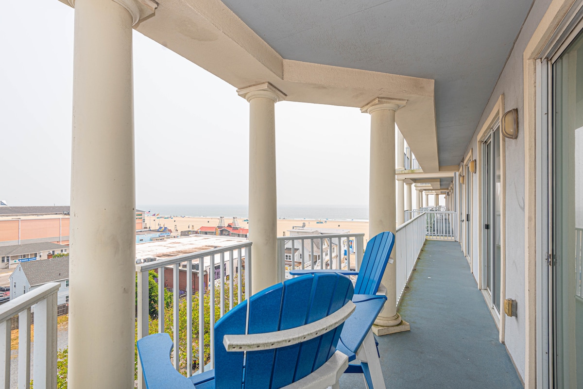 A spacious balcony features two blue adirondack chairs positioned near white columns. The railing offers unobstructed views of the ocean and beach area, with sand and distant umbrellas visible in the background. Soft lighting enhances the tranquil atmosphere of this outdoor space.