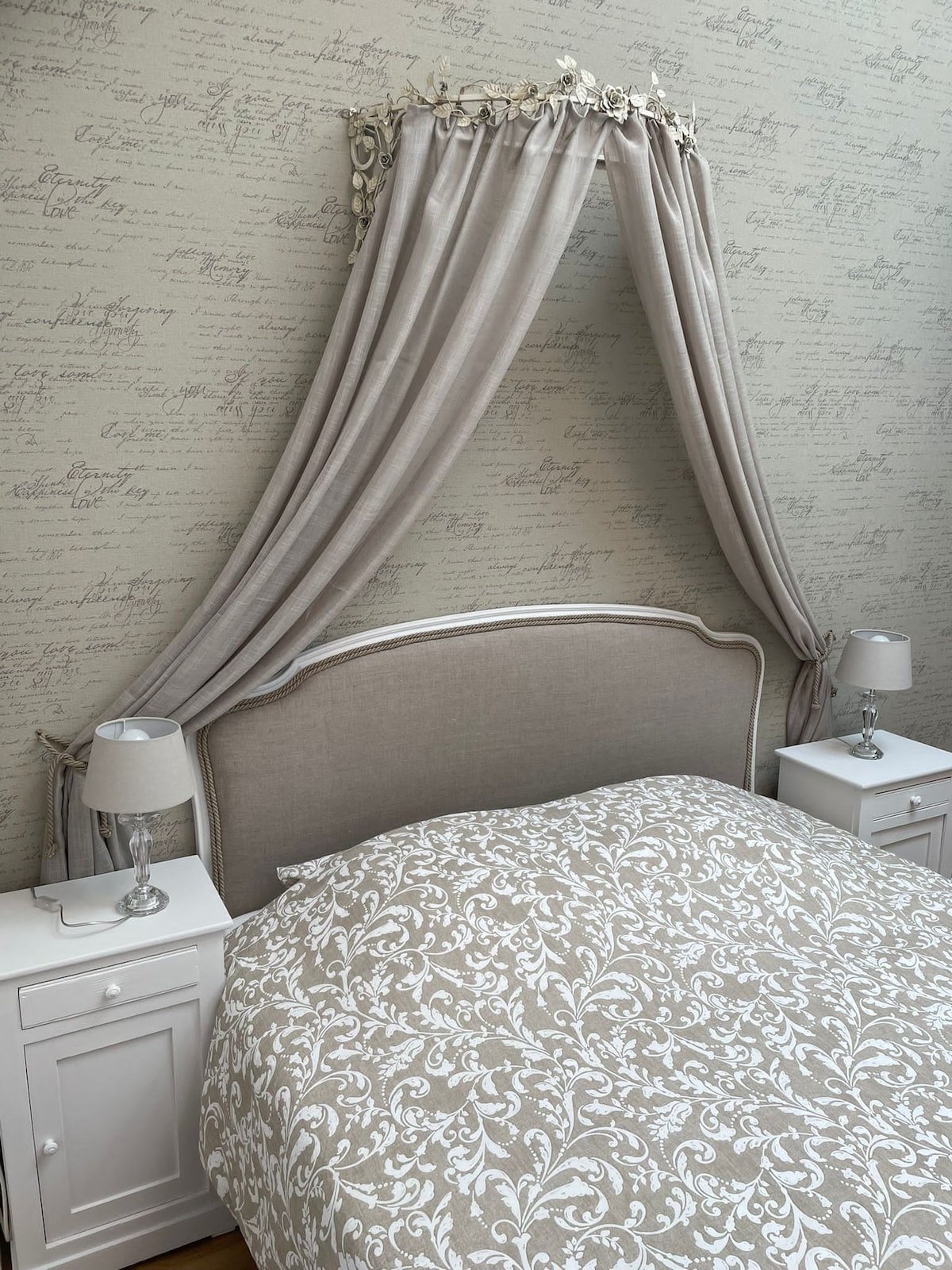 A cozy sleeping area features a spacious bed with a decorative coverlet adorned with a subtle floral pattern. Elegant bedside tables with lamps are positioned on either side of the headboard, which is framed by soft drapery that adds a romantic touch.