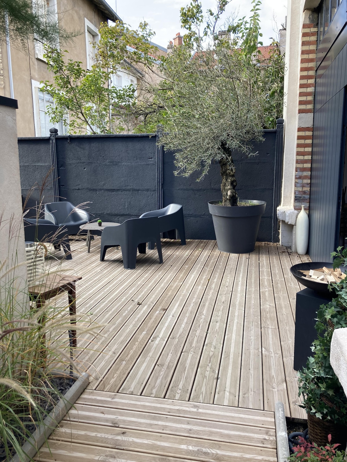 An outdoor terrace features a wooden deck, inviting relaxation with two modern chairs situated beside a large potted olive tree. The area is enclosed by a tall dark fence, creating a private atmosphere enhanced by nearby greenery.