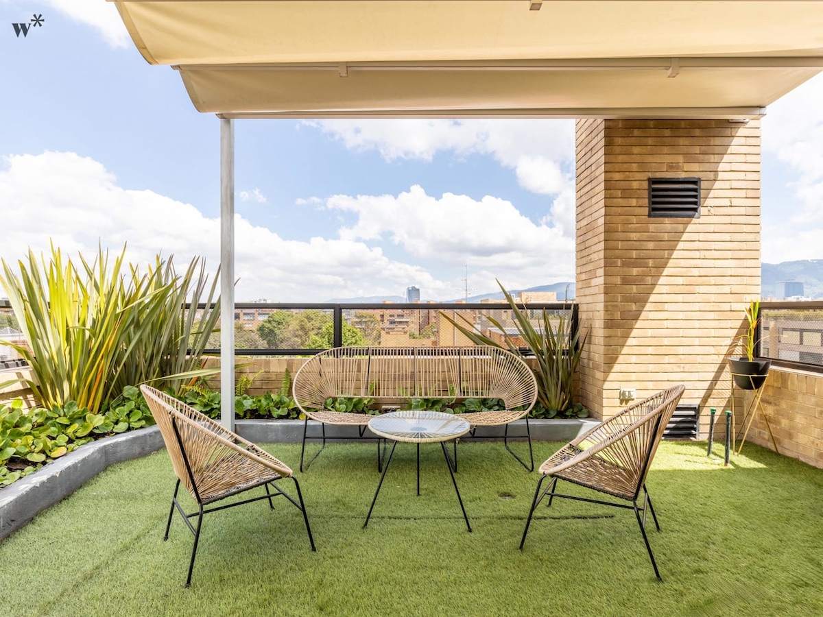 A private terrace is showcased, featuring four rattan chairs surrounding a circular table. Lush greenery lines the perimeter, complementing the modern brick structure. The open space is illuminated by natural light, with a clear blue sky and scattered clouds visible in the background.