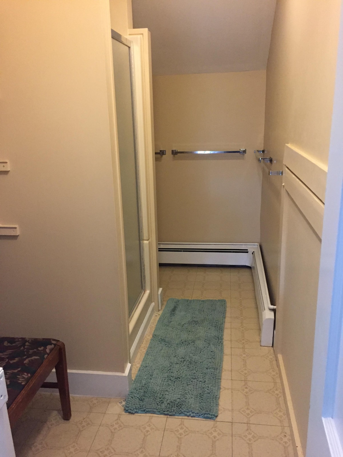 A bathroom is shown featuring a glass shower enclosure and a towel rack mounted on the wall. A light-colored rug is placed on the tiled floor, and a small wooden bench is positioned against the wall, enhancing the functional space.