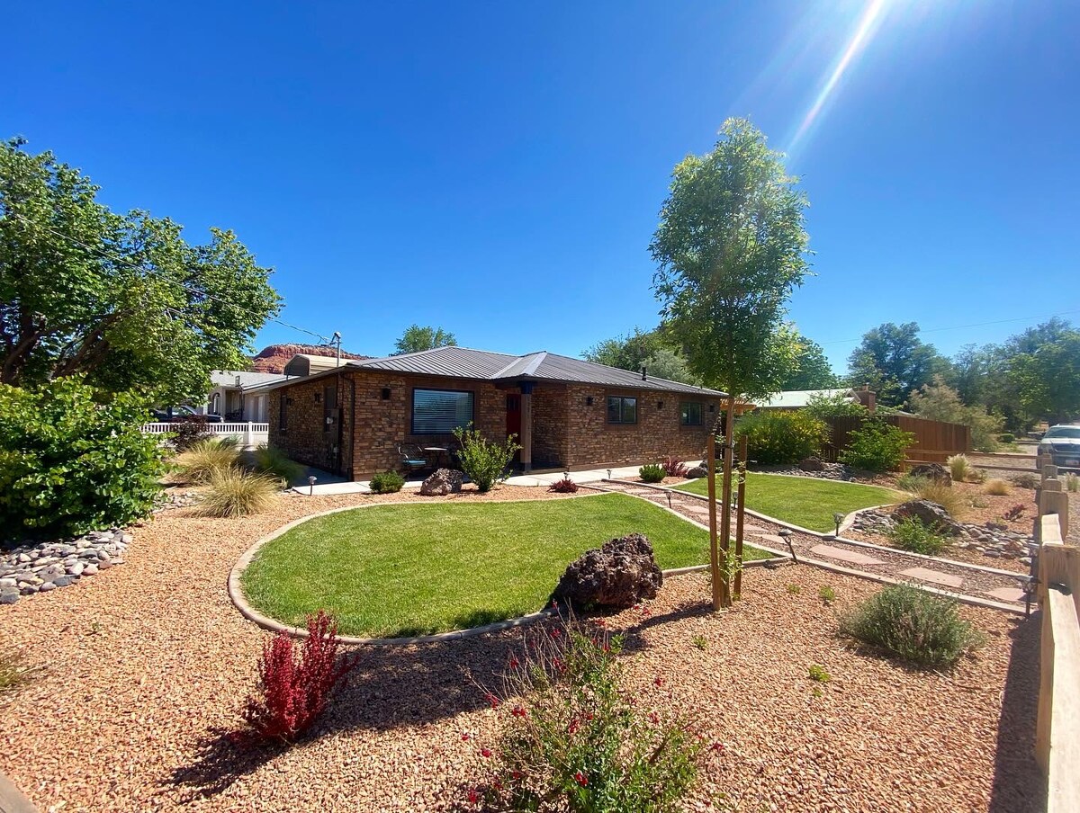 The exterior of a single-story home is framed by a landscaped yard, featuring a mix of grass, decorative rocks, and flowering plants. A pathway leads to the entrance, flanked by trees and shrubs under a clear blue sky.