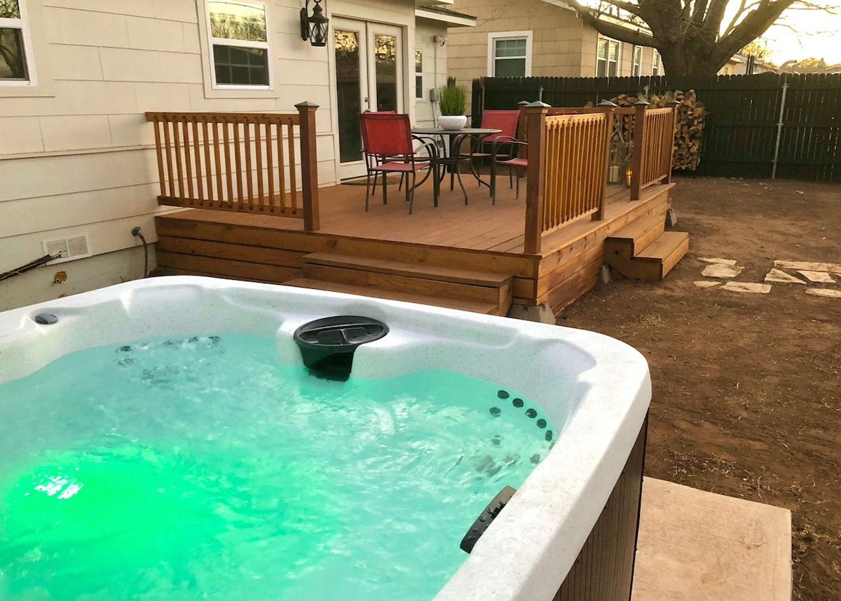 A hot tub is positioned in the foreground, showcasing bubbling water illuminated with changing colors. Behind, a wooden deck features a dining table and four chairs, surrounded by a spacious, fenced backyard that offers privacy and outdoor enjoyment.