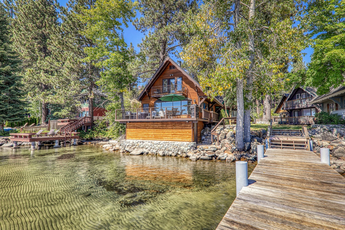 The cabin is positioned on the water's edge, surrounded by tall trees and a clear blue sky. A spacious deck overlooks the lake, while a wooden pier extends into the water, enhancing the serene lakeside setting.