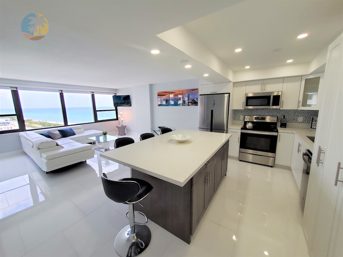 A spacious, modern kitchen with a large island and seating for four. The stainless steel appliances are complemented by sleek cabinetry. Natural light floods the open living area, showcasing ocean views through large windows. A comfortable seating area with a sectional couch is visible.