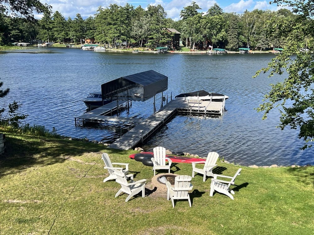 A serene outdoor area features a circular arrangement of Adirondack chairs surrounding a fire pit. A private dock extends into the calm water, providing access to a scenic waterfront. Lush greenery and trees frame the space, creating a welcoming environment for relaxation.