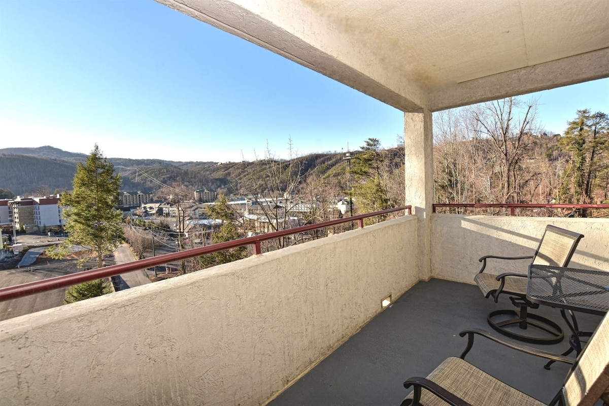 A covered deck offers views of distant mountains and the Gatlinburg landscape. Two stylish chairs are positioned on a concrete surface, complemented by a railing that adds to the space's openness. Natural light fills the area, enhancing the tranquil environment.