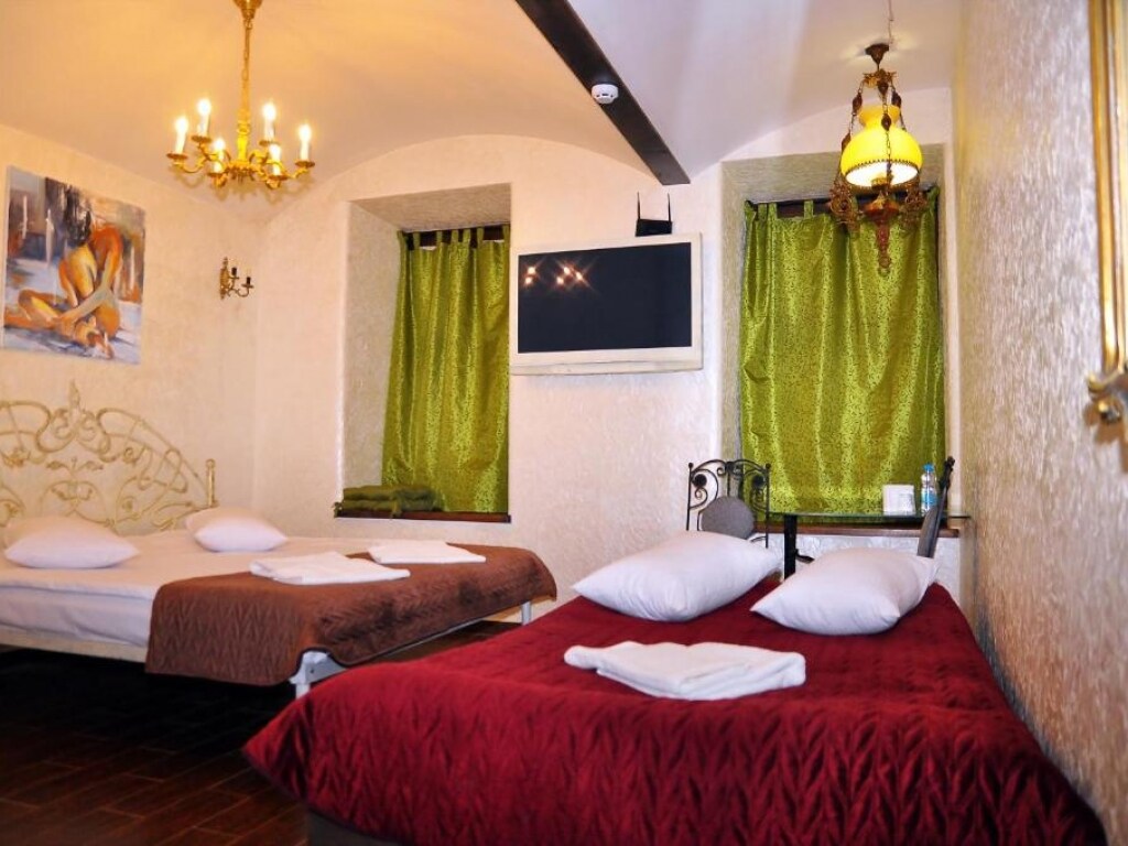 A cozy room features two beds with plush bedding and soft pillows. Bright green curtains frame the windows, allowing natural light to fill the space. A flat-screen TV is mounted on the wall, complementing the softly illuminated chandelier above.