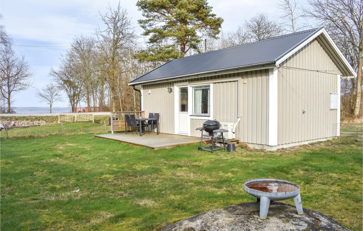 A light-colored cottage is surrounded by a grassy area, with a wooden deck featuring outdoor seating and a barbecue. Nearby, a fire pit is visible on a rocky surface. Trees provide a natural backdrop, and the proximity to water is suggested in the distance.