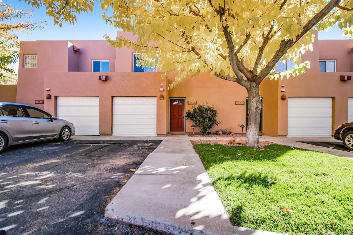 A Southwestern-style townhome is visible, featuring a welcoming entrance with a wooden door. Two garage doors are located on either side, and a well-manicured lawn is present in front, complemented by a large tree with golden leaves casting shade on the walkway.