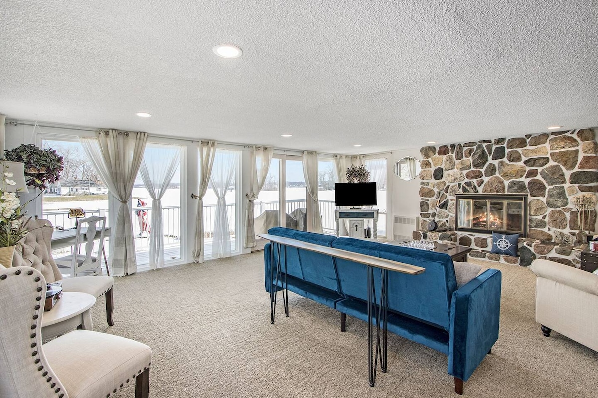 A spacious great room features a stone fireplace and ample natural light from large windows overlooking the lake. Comfortable seating options include a blue couch and an armchair. A dining space is visible nearby, enhancing the welcoming atmosphere.