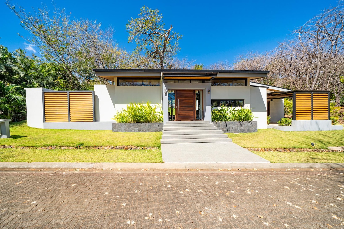 The modern villa is showcased from the front, featuring a sleek design with large windows and wooden accents. Steps lead up to a welcoming entryway, framed by lush green landscaping and a majestic Nya tree, highlighting the home's integration with its natural surroundings.