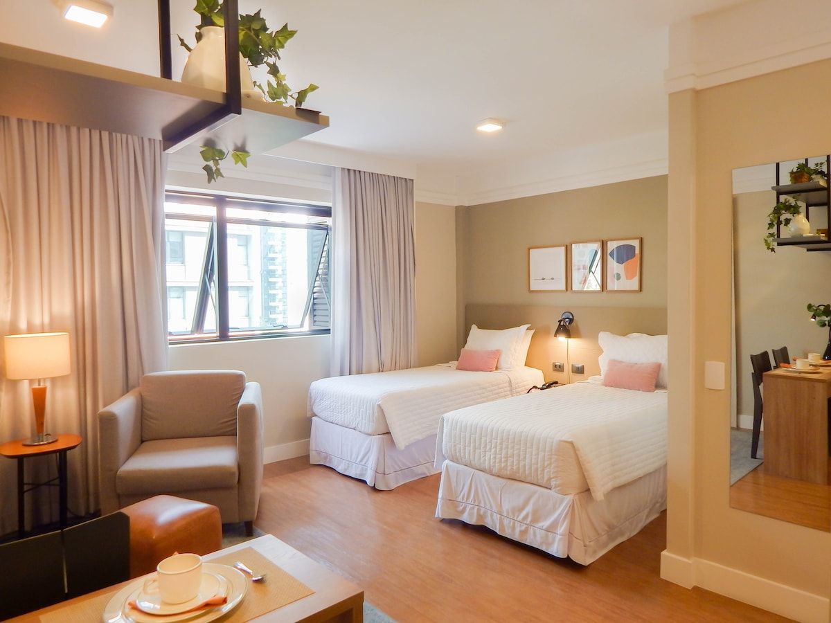 The cozy flat features two single beds adorned with crisp white linens. A comfortable armchair sits beside a lamp, while a sleek table is positioned nearby. Natural light enters through a large window, complemented by light gray curtains.