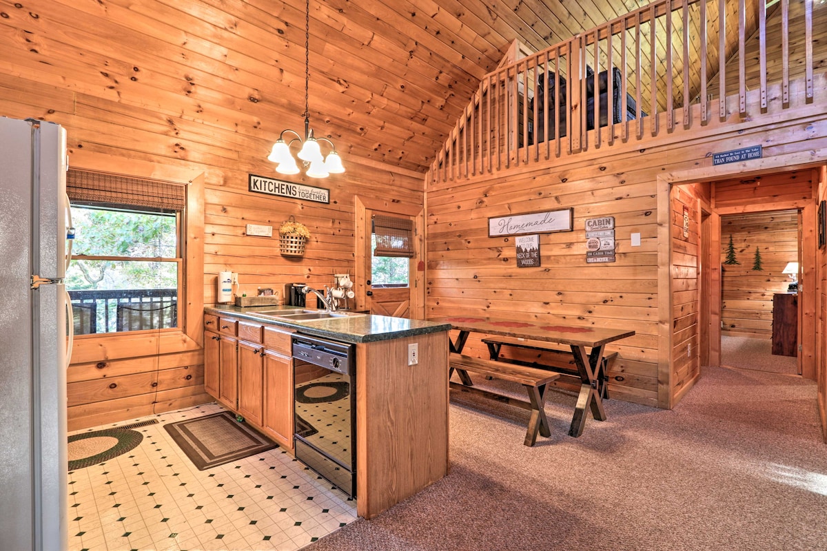 The kitchen area features wooden walls and high ceilings, creating a rustic ambiance. A spacious granite countertop is paired with wooden cabinetry, while a long picnic-style table offers ample seating. Large windows provide natural light and views of the surrounding trees.