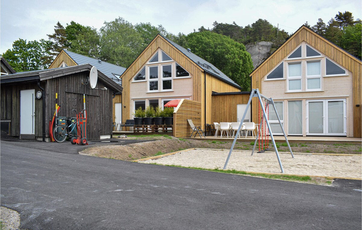 Two modern vacation homes are positioned alongside each other, featuring large windows and distinct triangular roofs. A sandy area for play is visible in the foreground, equipped with a swing set. Nearby, bicycles are parked against a wooden shed, emphasizing a family-friendly environment.