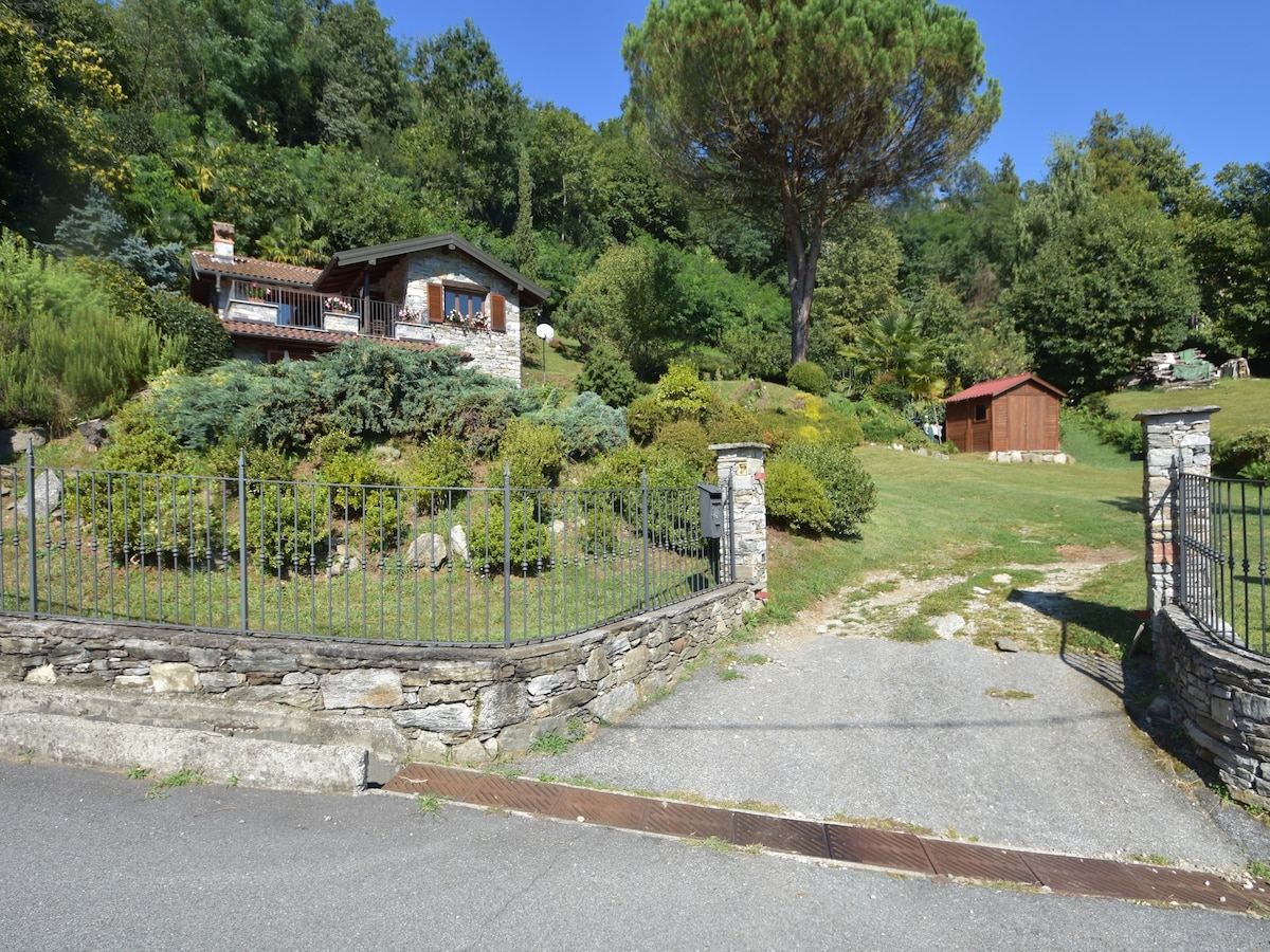 A welcoming entrance to the apartment features a stone wall and a wrought iron gate. The landscaped garden surrounds the property, showcasing a variety of greenery and trees. A charming wooden structure can be seen in the background, enhancing the serene outdoor environment.