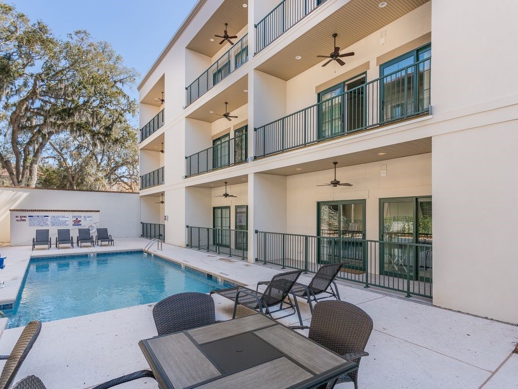 An outdoor swimming pool is situated in a sunny area, surrounded by lounge chairs and small tables. The three-story building features multiple balconies with green railings, providing a view of the pool area below. Trees are visible in the background.