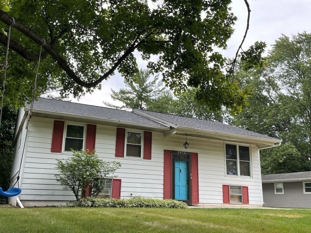 A cozy split-level home is framed by green trees and grass, featuring a blue front door and red shutters. A swing hangs from a nearby tree, adding to the inviting outdoor space.