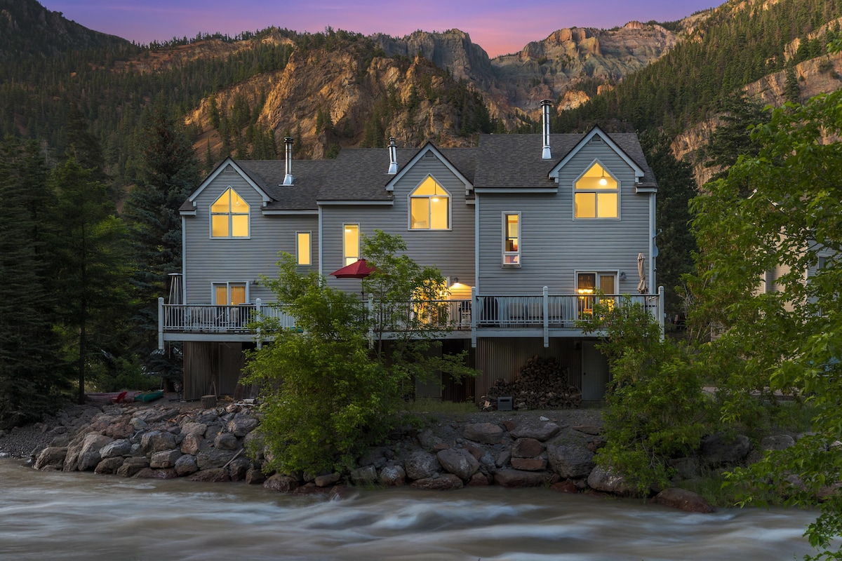 The townhome is situated alongside the Uncompahgre River, surrounded by lush greenery and rocky terrain. Its exterior showcases multiple large windows, reflecting warm light from within. The backdrop features majestic mountains, enhancing the serene atmosphere as the evening sky transitions to twilight.